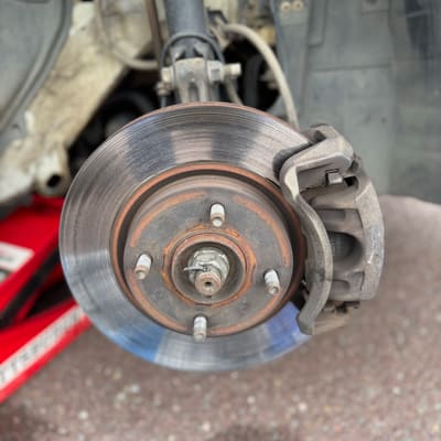 Worn front brake rotor still on the car, its face scored and the outer edge ridged