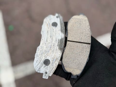 Gloved hand holding two worn brake pads: one gouged through the friction material, the other cracked across the middle